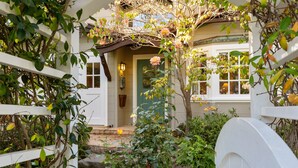 Um portão de madeira branca dá acesso a uma casa com porta verde, cercada por vegetação exuberante e plantas floridas.