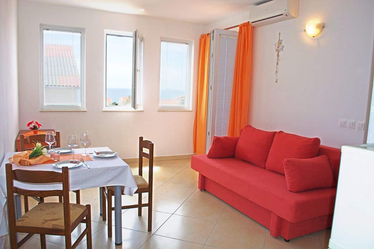 A dining area with a table set for four, two windows with blinds, a red sofa, and a wall-mounted air conditioner.