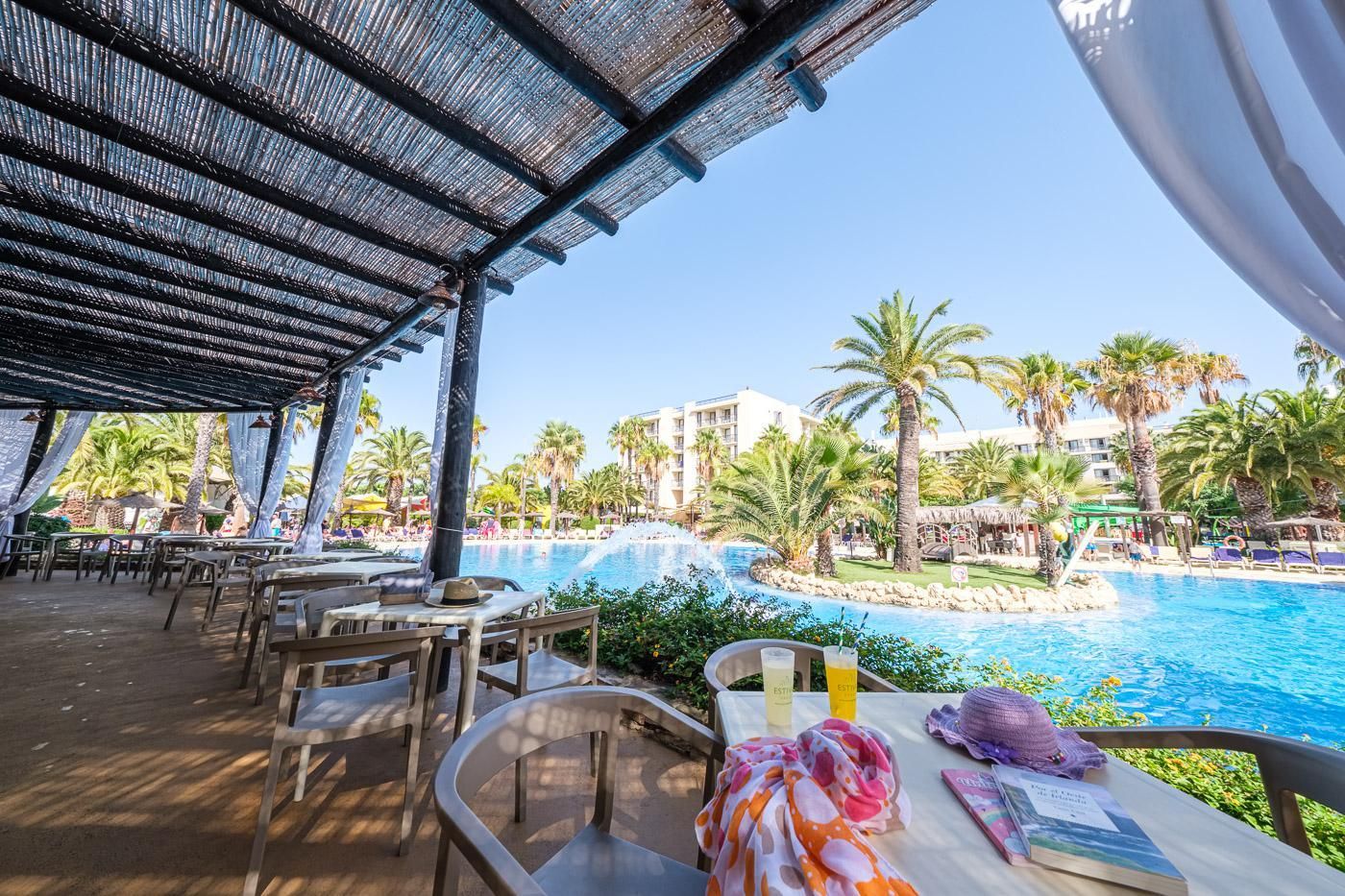 An outdoor dining area with tables and chairs, overlooking a pool with palm trees and a building in the background.
