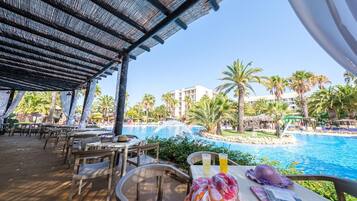 An outdoor dining area with tables and chairs, overlooking a pool with palm trees and a building in the background.