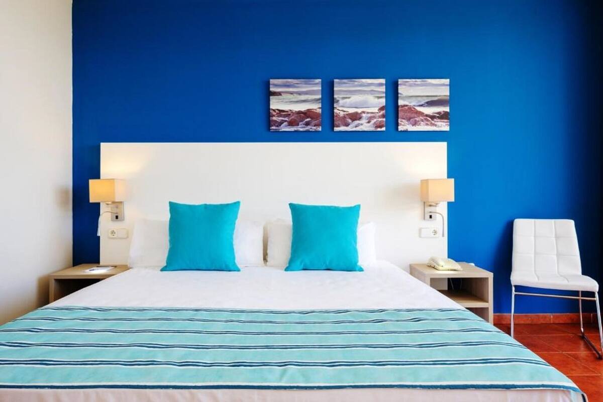 Ein Hotelzimmer mit einem großen Bett, zwei blauen Kissen, einem weißen Kopfteil, zwei Nachttischlampen, einem weißen Sessel und zwei gerahmten Bildern an der Wand.