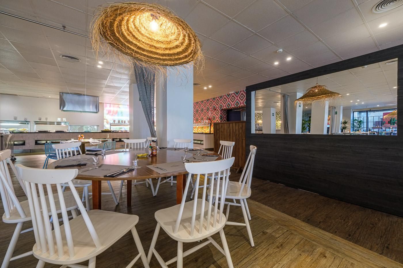 A modern dining area with white chairs and a wooden table, a large woven light fixture, and a view of the kitchen area in the background.