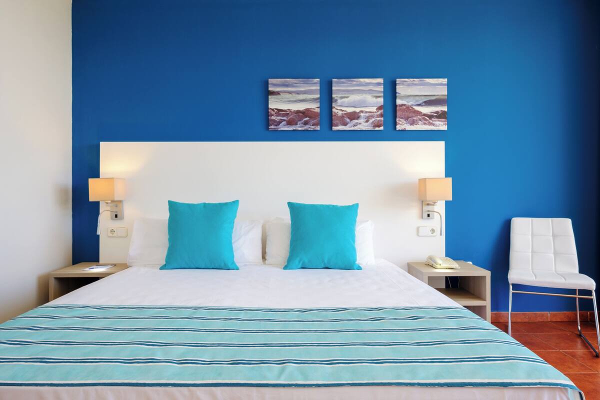 Ein Hotelzimmer mit einem großen Bett, zwei Nachttischen, einem weißen Stuhl und einer blauen Wand.