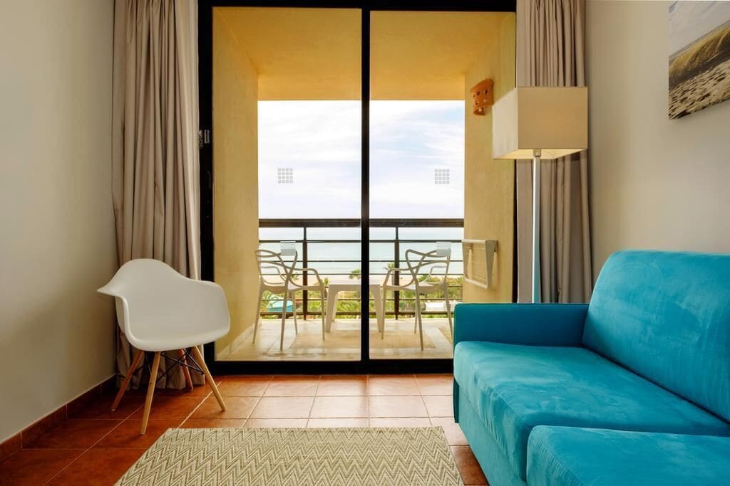 Ein Zimmer mit einem blauen Sofa, einem weißen Sessel, einer Stehlampe und Meerblick durch eine Schiebetür.
