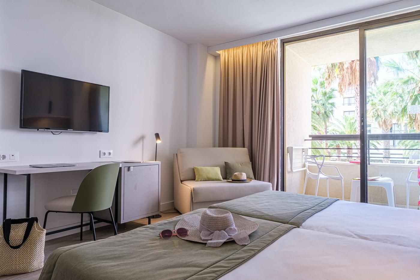 Quarto de hotel com cama, escrivaninha com computador, televisão, cadeira e varanda com vista para palmeiras.
