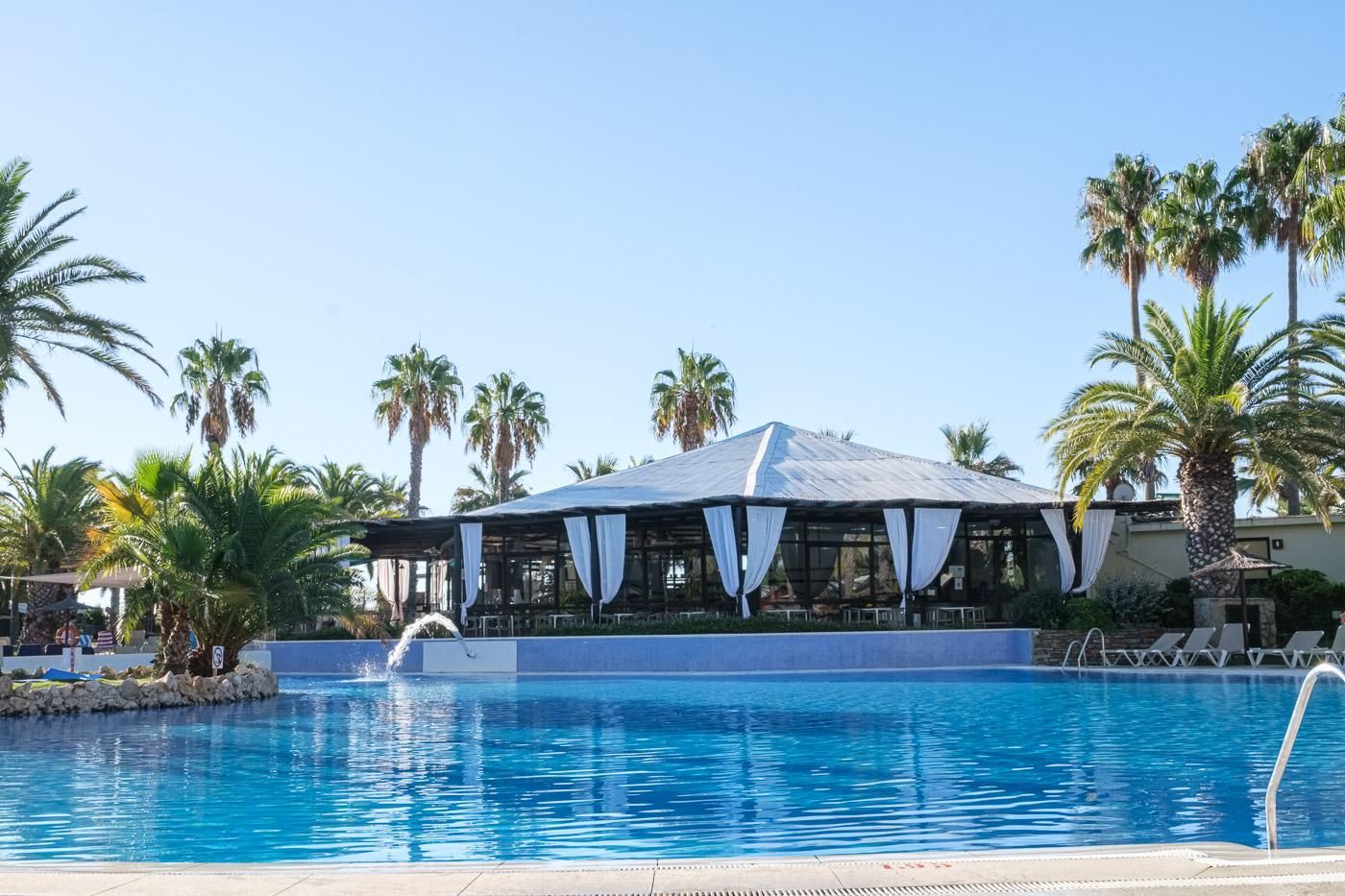 A pool area with a covered seating area, palm trees, and a clear blue sky.