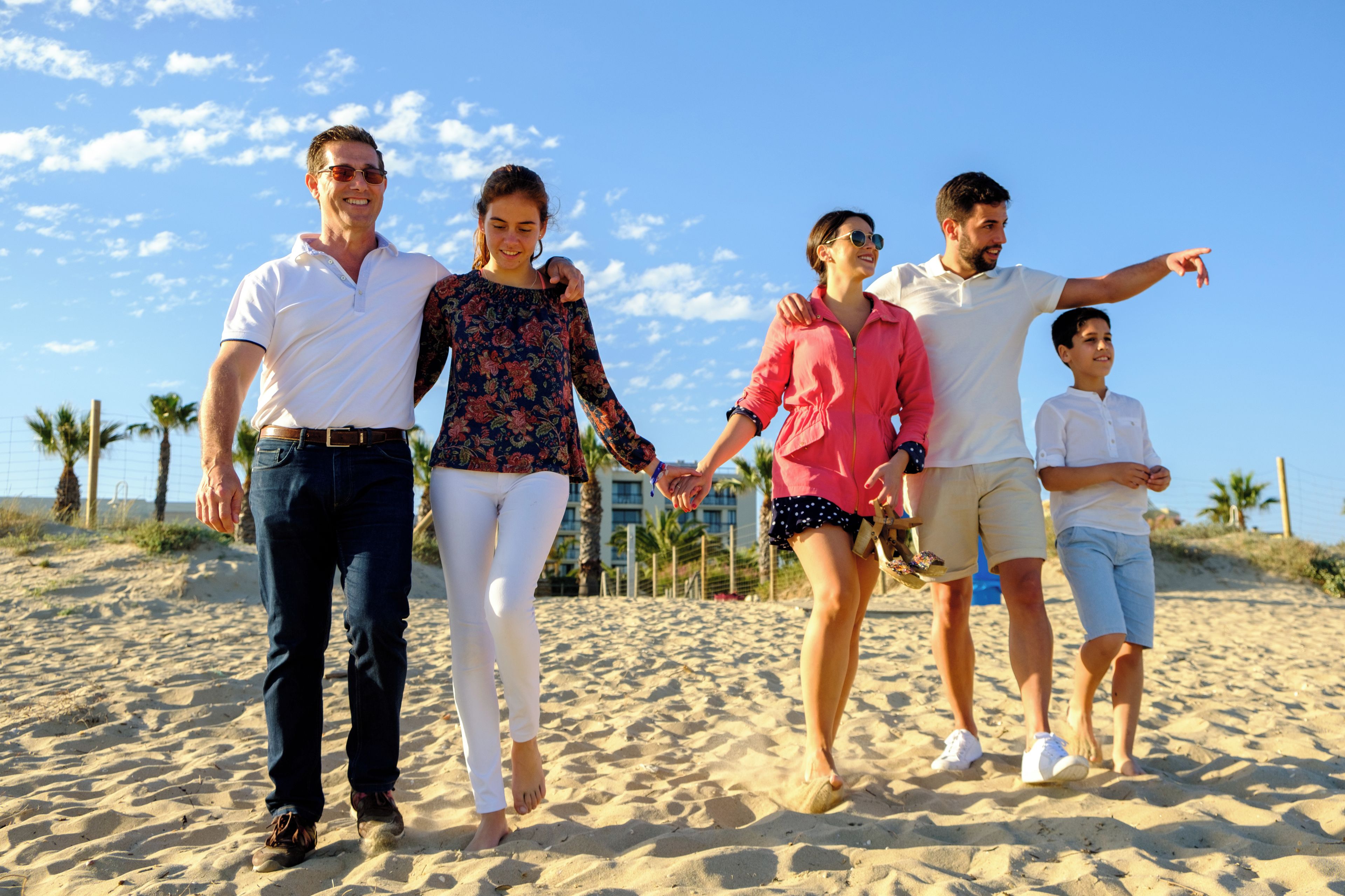 Una familia caminando sobre una playa arenosa, con palmeras y edificios al fondo.