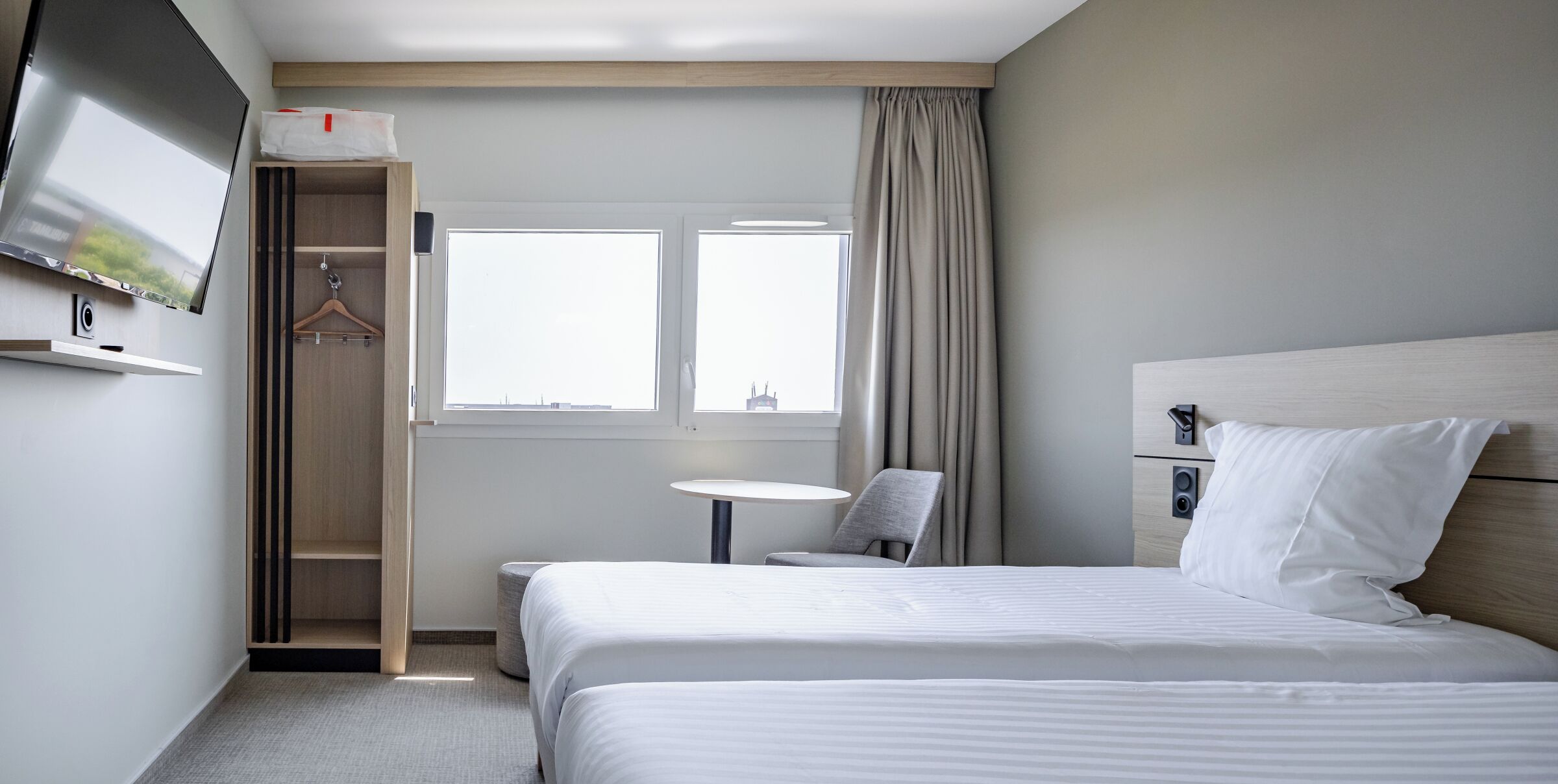 A hotel room with a bed, a TV mounted on the wall, a small table, a chair, and a window with curtains.