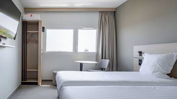 A hotel room with a bed, a TV mounted on the wall, a small table, a chair, and a window with curtains.