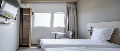 A hotel room with a bed, a TV mounted on the wall, a small table, a chair, and a window with curtains.