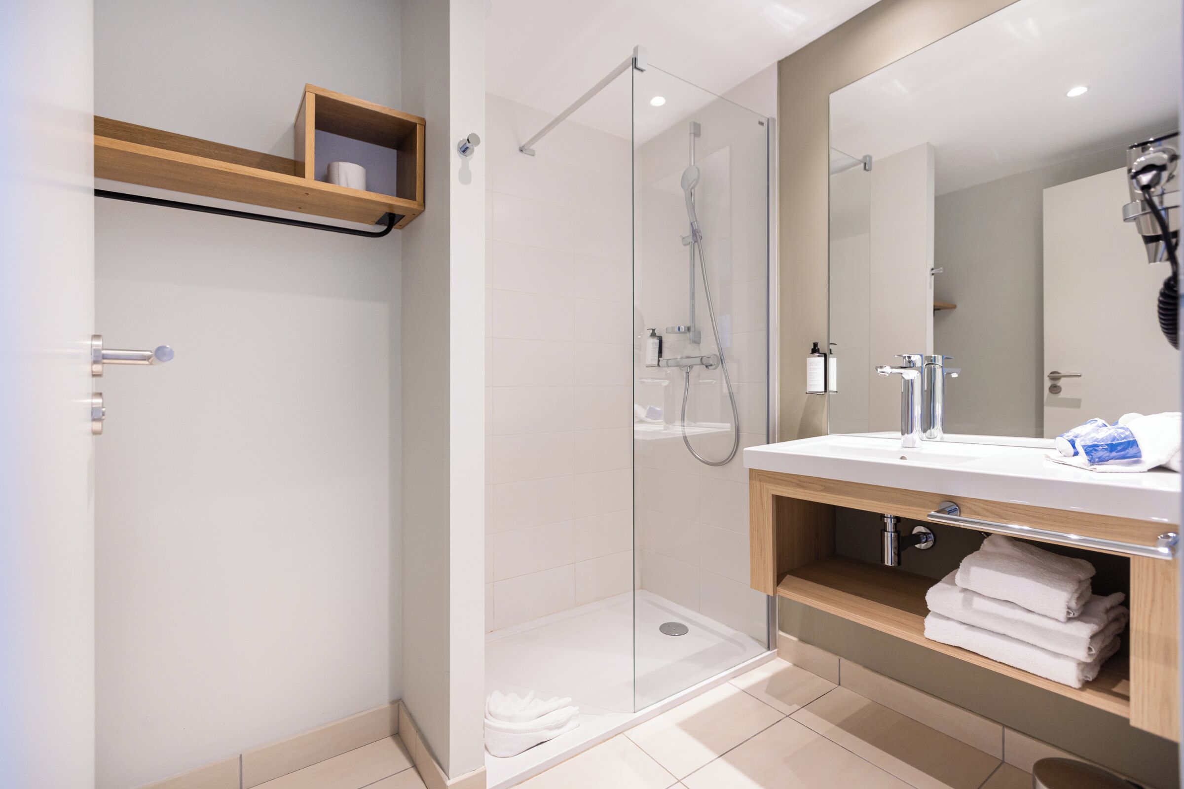 Un baño moderno con ducha envuelta en vidrio, un estante de madera con artículos de aseo y un espejo grande sobre el lavamanos.