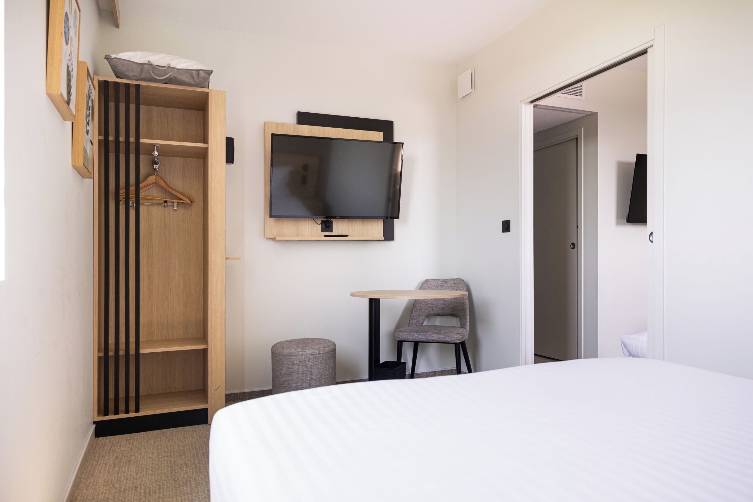 A hotel room with a bed, a TV mounted on the wall, a wooden wardrobe, a small table with a chair, and a door leading to another room.