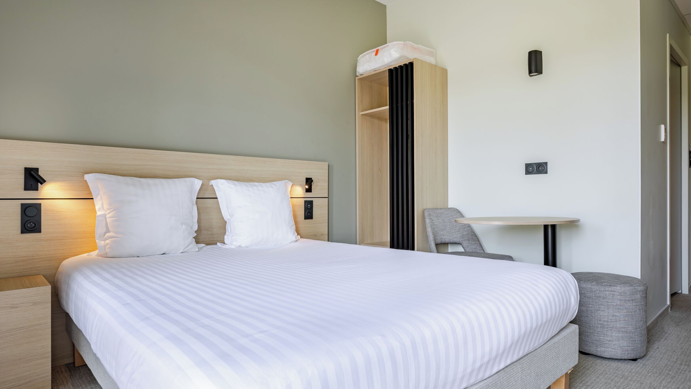 A neatly made bed with two pillows, a bedside table, a chair, and a small ottoman.