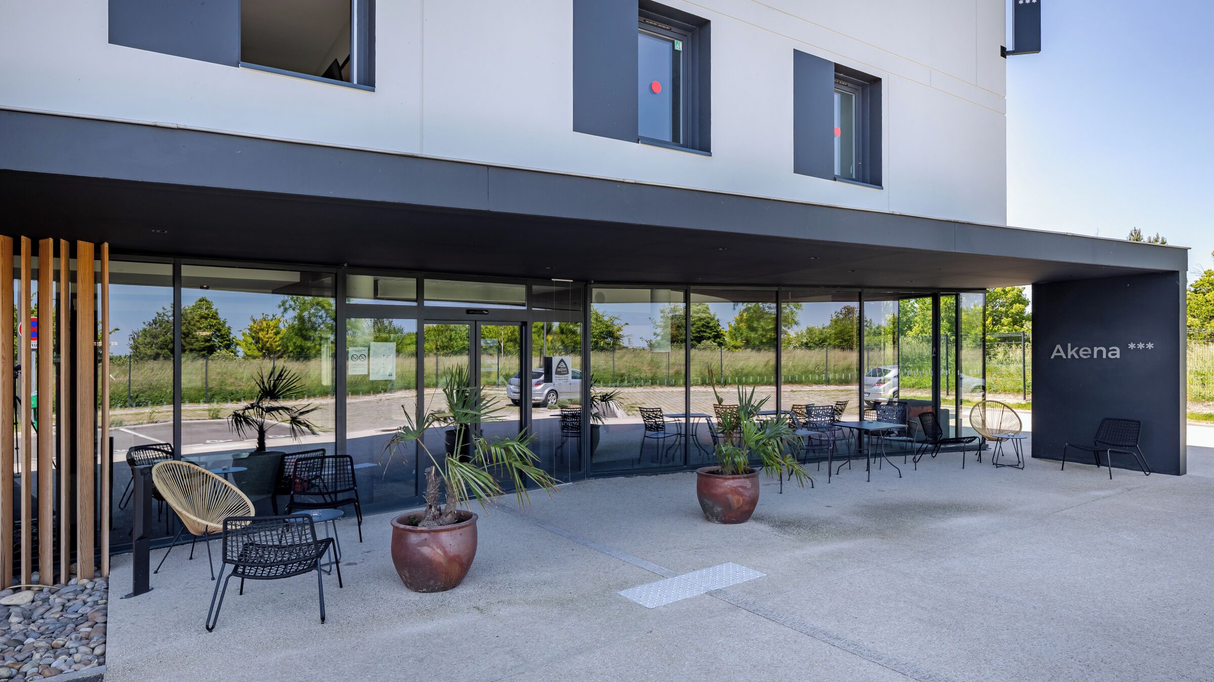 A modern building with a covered outdoor seating area, large windows, and a sign reading 'Akena ***'.