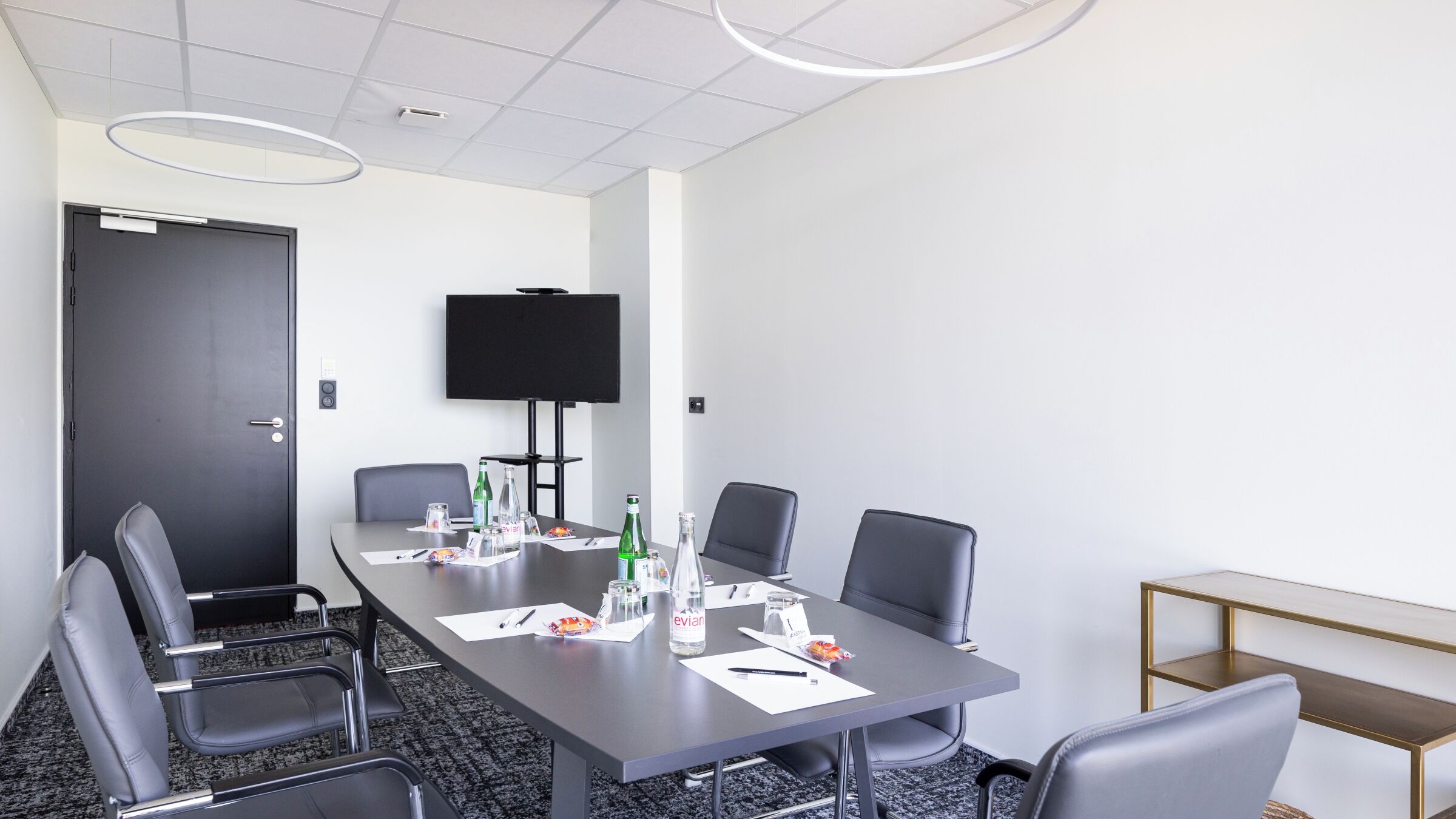 A conference room with a long table, six chairs, a television on the wall, and various items on the table.