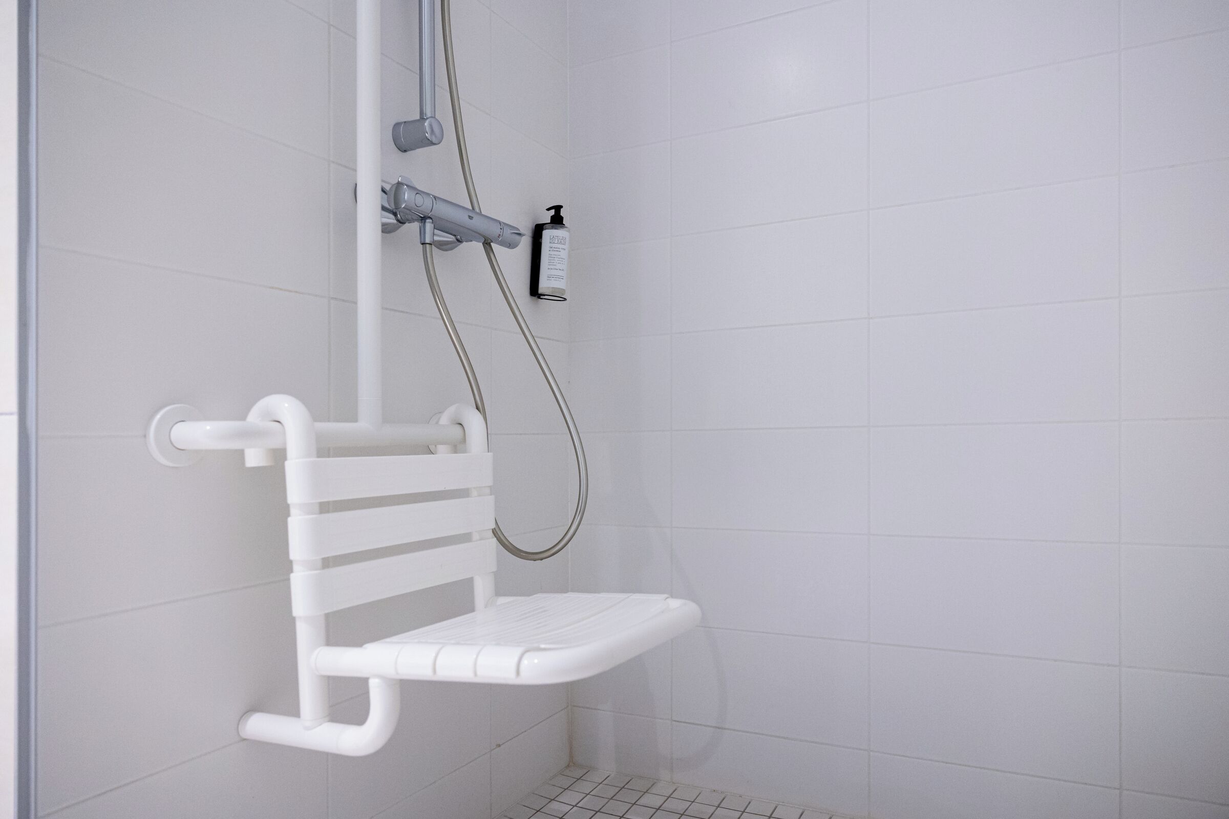 Ducha con una barra de apoyo blanca, un cabezal de ducha de mano y una botella de champú en la pared.