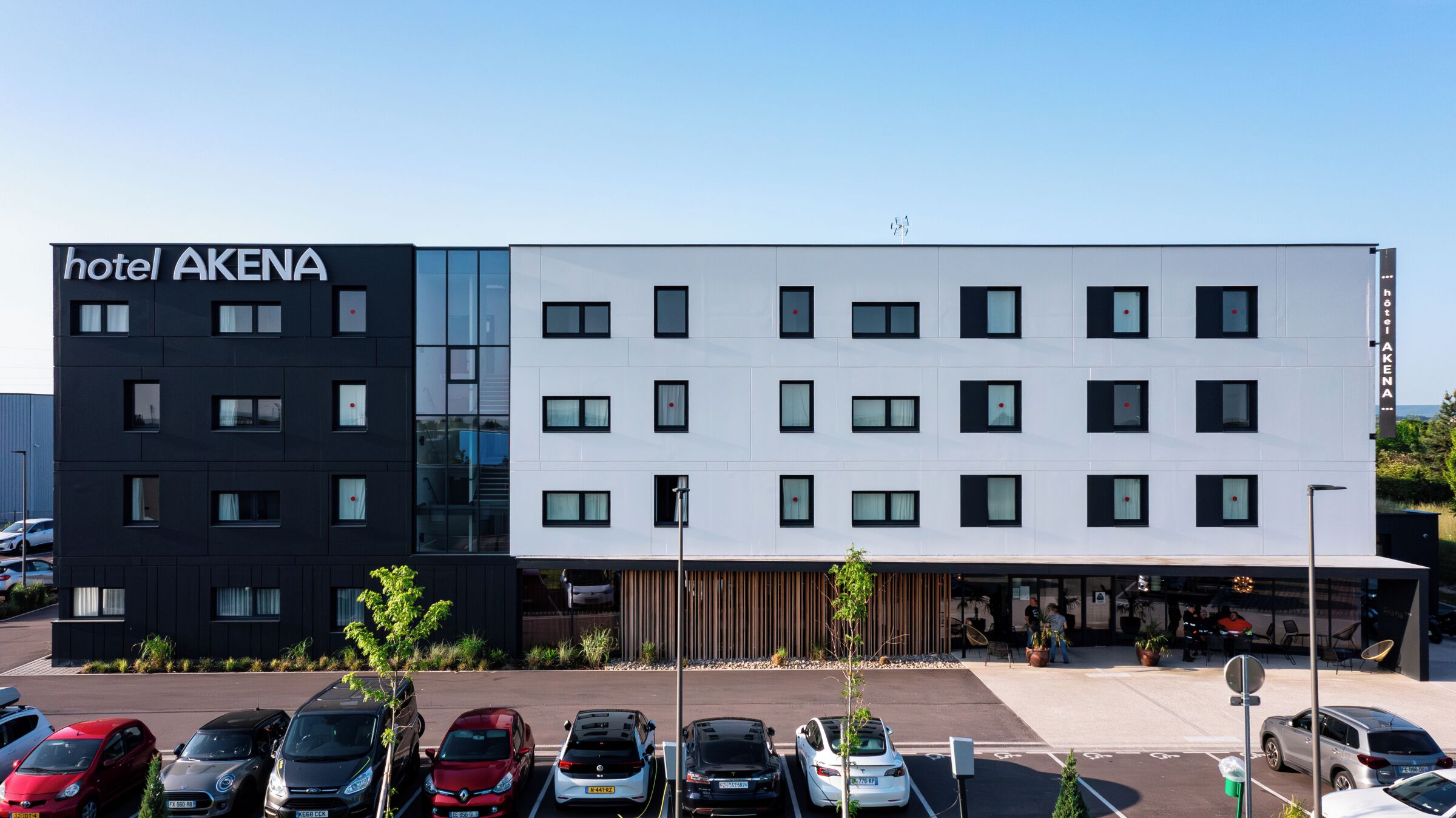 Een modern hotelgebouw met een tweetonig gevelontwerp, een parkeerplaats met diverse auto's en een heldere lucht.