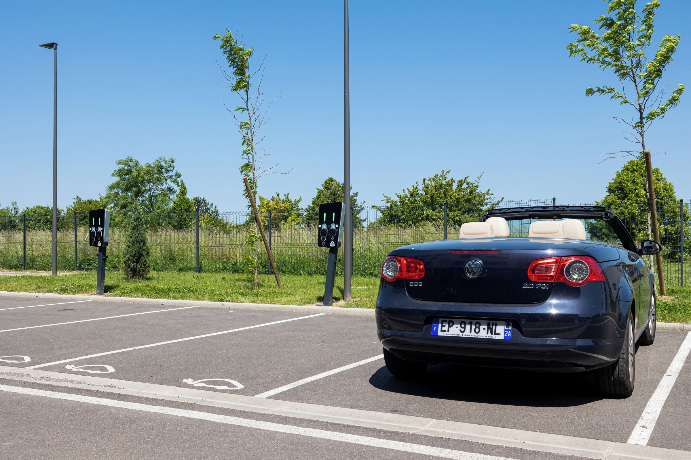 A parking lot with a blue convertible car, a pedestrian crossing, and a fence with trees.