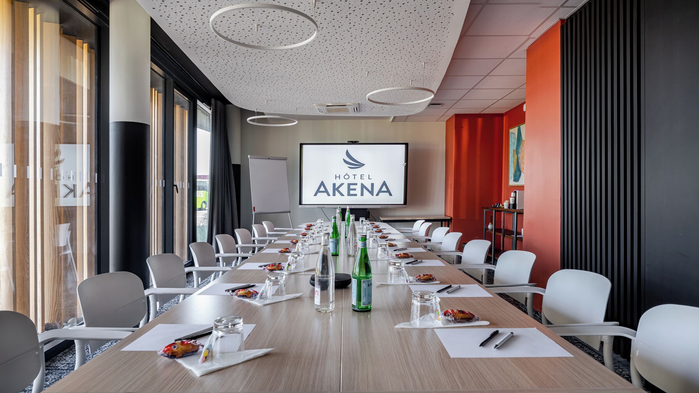 Ein Hotelkonferenzraum mit einem langen Tisch, weißen Stühlen und einer großen Leinwand, die den Namen des Hotels zeigt.