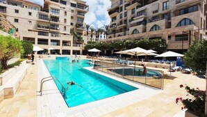 A swimming pool area with multiple levels of residential buildings, outdoor seating, and palm trees. - MAMAD - Deluxe 2BD Jaffa Dreams W Pool & Parking (Tel Aviv)