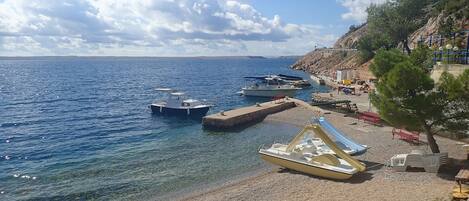 A pebbled beach with boats docked, a clear sky, and a rocky hillside with a tree.