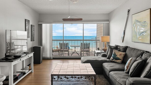 A living room with a view of the ocean, a ceiling fan, a TV, and a sofa with decorative pillows. - Seaside Beach House 203 · Top Floor · 14-Ft Balcony · Pool (Holmes Beach)