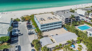An aerial view of a coastal hotel with a parking lot, a swimming pool, and a beach in the background. - Seaside Beach House 203 · Top Floor · 14-Ft Balcony · Pool (Holmes Beach)