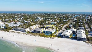 An aerial view of a coastal residential area with beachfront properties, including a hotel with a large building and multiple units, surrounded by greenery and a sandy beach. - Seaside Beach House 203 · Top Floor · 14-Ft Balcony · Pool (Holmes Beach)
