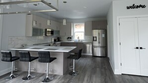 Een moderne keuken met een centraal eiland, barkrukken, roestvrijstalen apparatuur en een bordje met de tekst 'welkom' aan de muur.