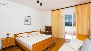 A bedroom with a wooden bed, a nightstand, a bedside lamp, a framed picture on the wall, and a view of a balcony with yellow curtains. - Guest House Ljubica (Dubrovnik)