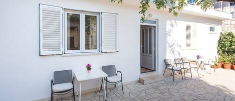 A patio area with a table and chairs, a white wall with shutters, and a stone floor.