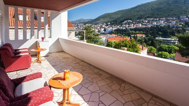 A balcony with red patterned sofas, a small round table with a drink, and a view of a town and hills. - Guest House Ljubica (Dubrovnik)