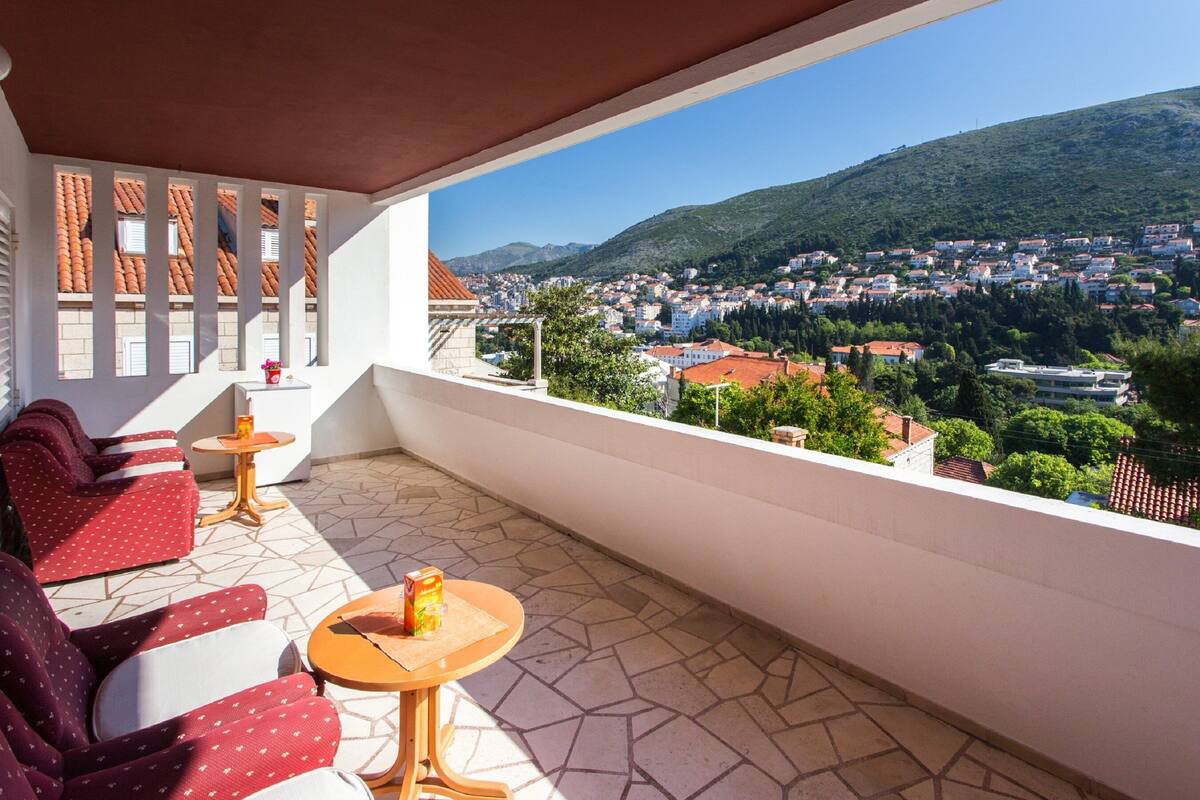 Un balcon avec des canapés à motifs rouges, une petite table ronde sur laquelle se trouve une boisson, et une vue sur une ville et des collines.