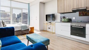 A modern apartment with a large window, a sofa, a small table, a kitchen with a Bosch oven, and a television. - Backup-Power Lavish Eclectic City View Apartment on Popular Bree Street (Cape Town)