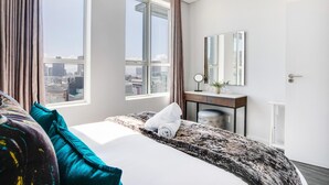 A hotel room with a large bed, a desk with a mirror, and a view of the city through large windows.