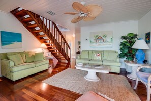A living room with a wooden staircase, two green sofas, a coffee table, a fan, and a painting of a house. - Manatee Lookout (Anna Maria)