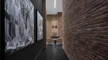 A hallway with a brick wall, a black tiled floor, and a framed artwork depicting a car and buildings. There is a small table with chairs in the distance.