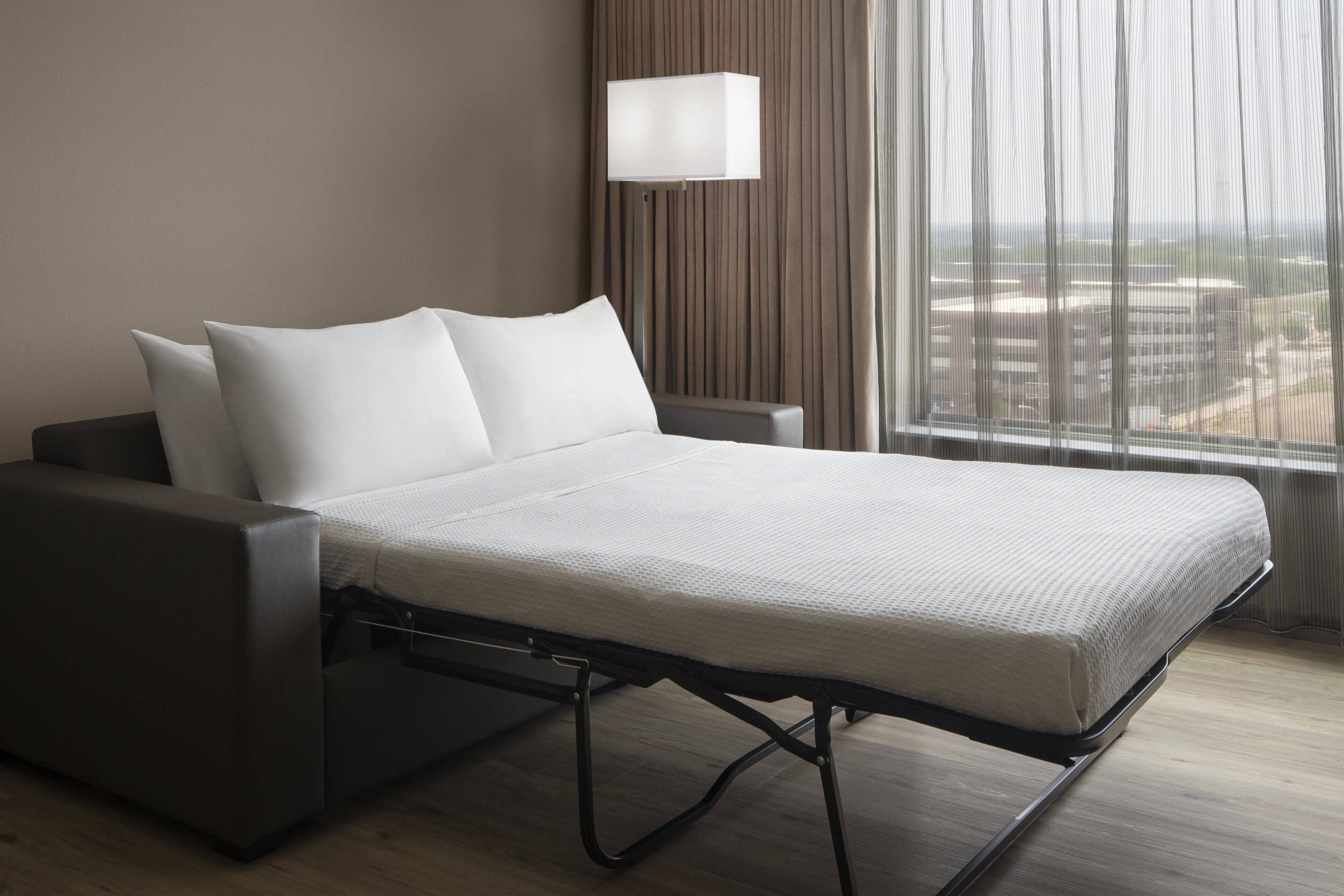 A modern hotel room with a bed, a bedside table, a lamp, and a view of the city through large windows.