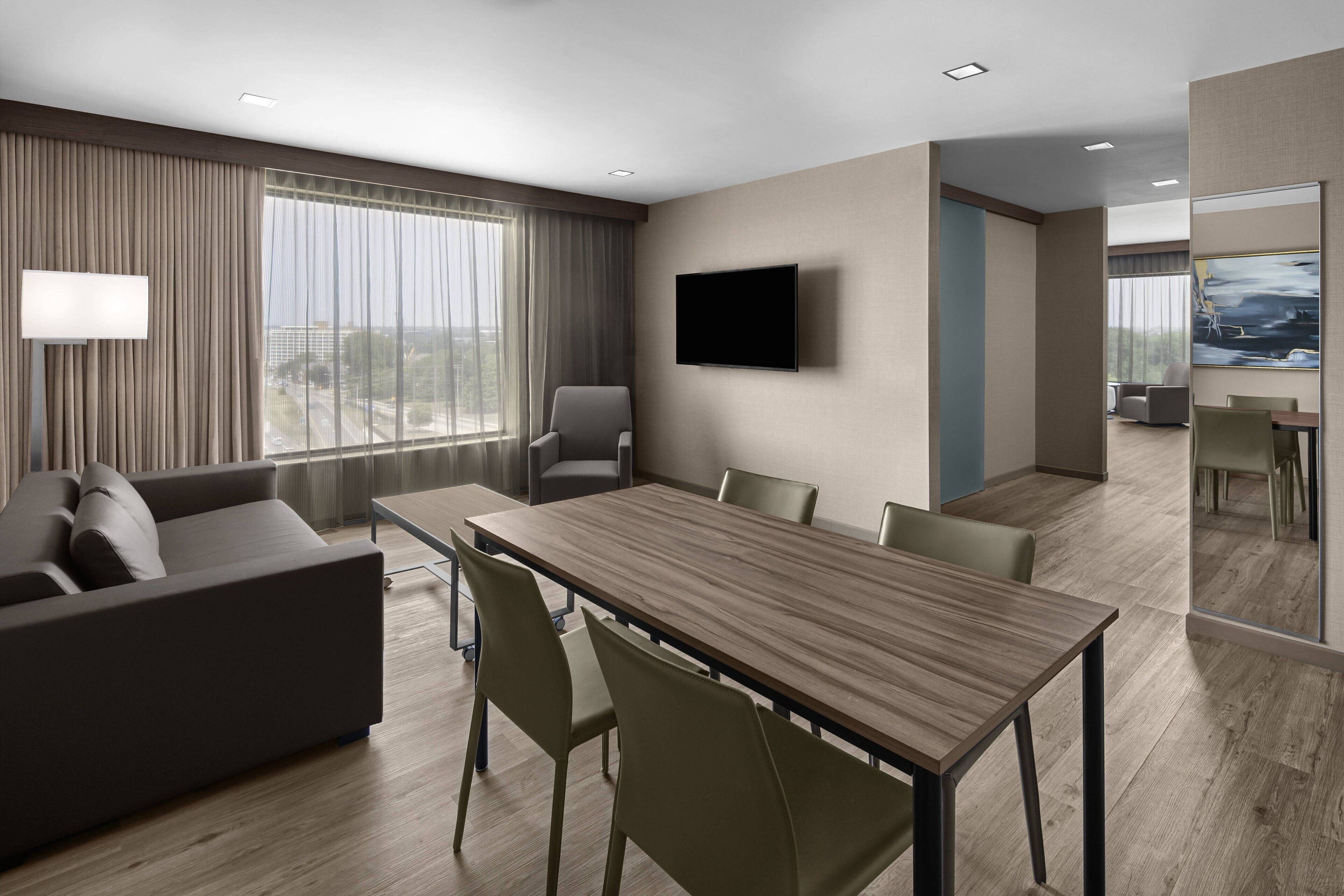 Une chambre d’hôtel moderne dotée d’une grande table à manger, de sièges confortables, d’un téléviseur à écran plat et de grandes fenêtres offrant une vue sur la ville.