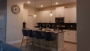 A modern kitchen with white cabinets, a central island with bar stools, a clock on the wall, and pendant lighting. - 801DSD: Stunning 5 Bedroom Villa - Private Pool and Spa with Nature View (Kissimmee)