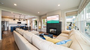 A spacious living room with a large sofa, a flat-screen TV mounted above a fireplace, and a kitchen in the background. - Welcome to Don’t Stop Believin’ at Lake Chelan (Chelan)
