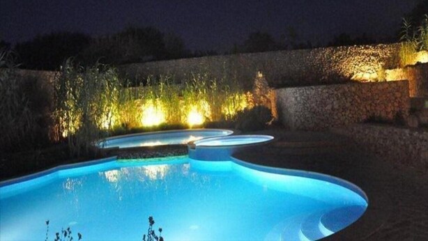 A swimming pool with a curved design, surrounded by a stone wall and illuminated plants. - Misrah Simar (Qala)