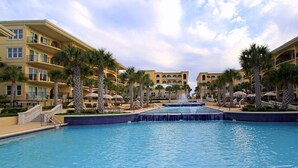 Ein Hotelpool mit einem zentralen Wasserspiel, umgeben von Palmen und einem mehrstöckigen Gebäude mit Balkonen.