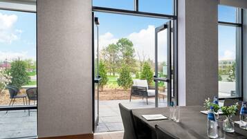 A modern hotel room with large windows, a dining table set for a meeting, and a view of the outdoor patio.