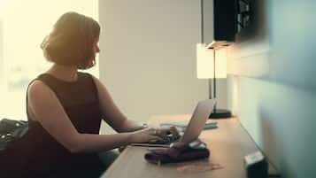 A person working on a laptop at a wooden desk with a lamp and a notebook.