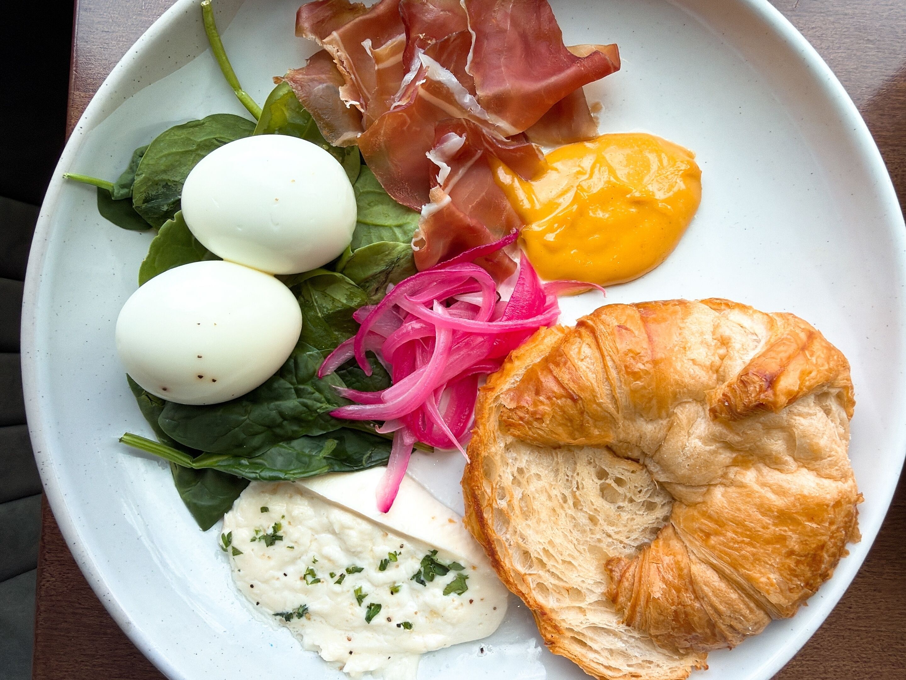 A plate with eggs, spinach, prosciutto, pickled onions, a croissant, and a creamy sauce.