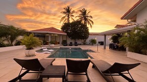 Ein Poolbereich mit Liegestühlen und einer überdachten Sitzgelegenheit, umgeben von Palmen und einem Sonnenuntergangshimmel.