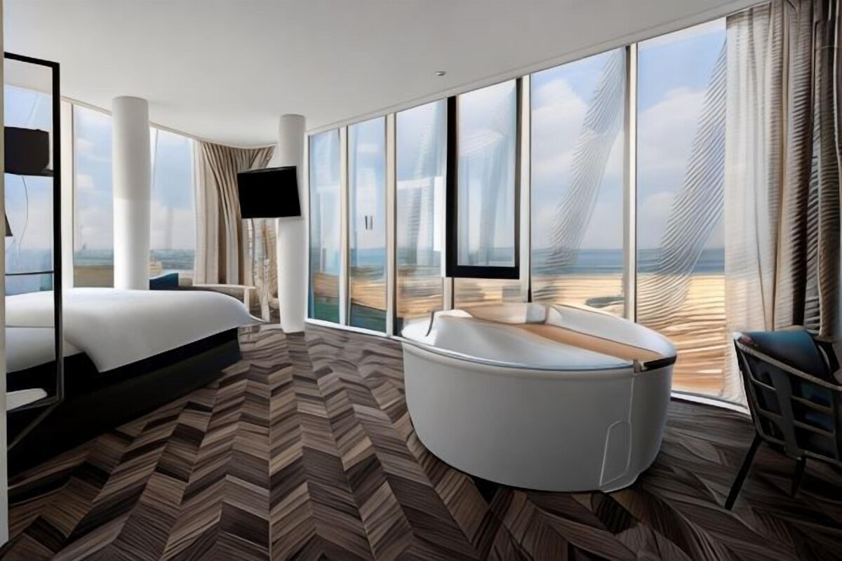 A modern hotel room with a large bed, a curved seating area, and a view of the beach.