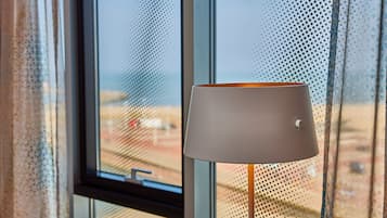 Una lámpara con pantalla blanca y un interruptor, ubicada cerca de una ventana con vista a la playa.
