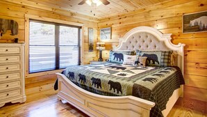Ein Schlafzimmer einer Holzhütte mit Bett, Schminktisch, Deckenventilator und einem Fenster mit Jalousien.